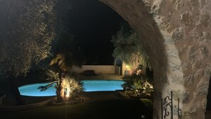 Vista nocturna de una zona de piscina con una estructura de piedra en arco, plantas iluminadas y cielo despejado.