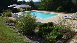 A rectangular swimming pool with two umbrellas, surrounded by a tiled deck and lounge chairs.