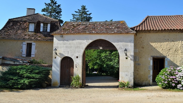 A rustic stone building with a large arched entrance, a small garden with flowers, and a clear blue sky.