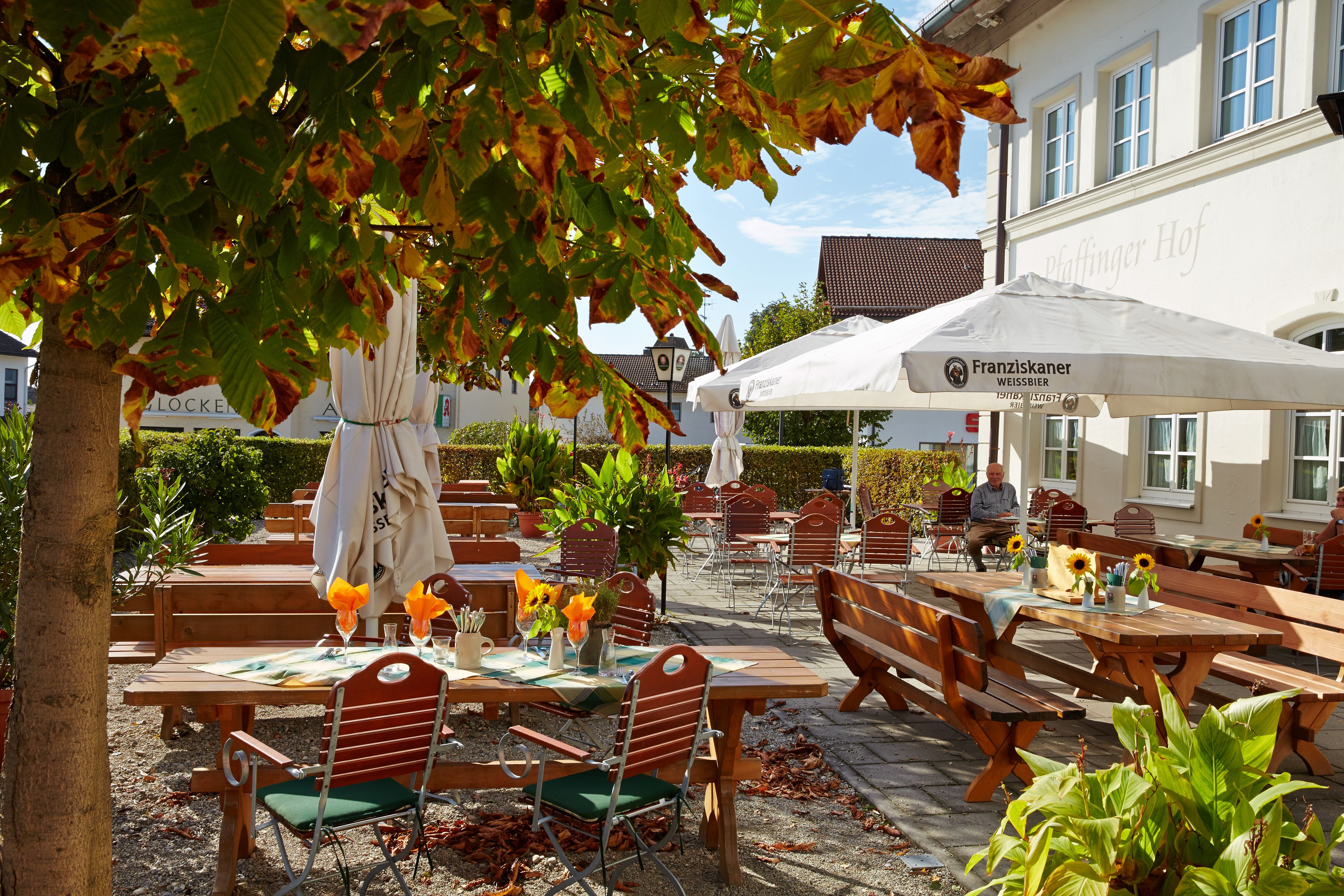 Un espace de café en plein air avec des tables et des chaises en bois, un parasol blanc et un bâtiment en arrière-plan.