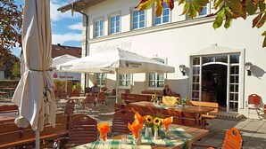 Un espace extérieur aménagé avec des tables et des chaises, un parasol blanc et un bâtiment portant l’enseigne « Paffinauer Hof ».