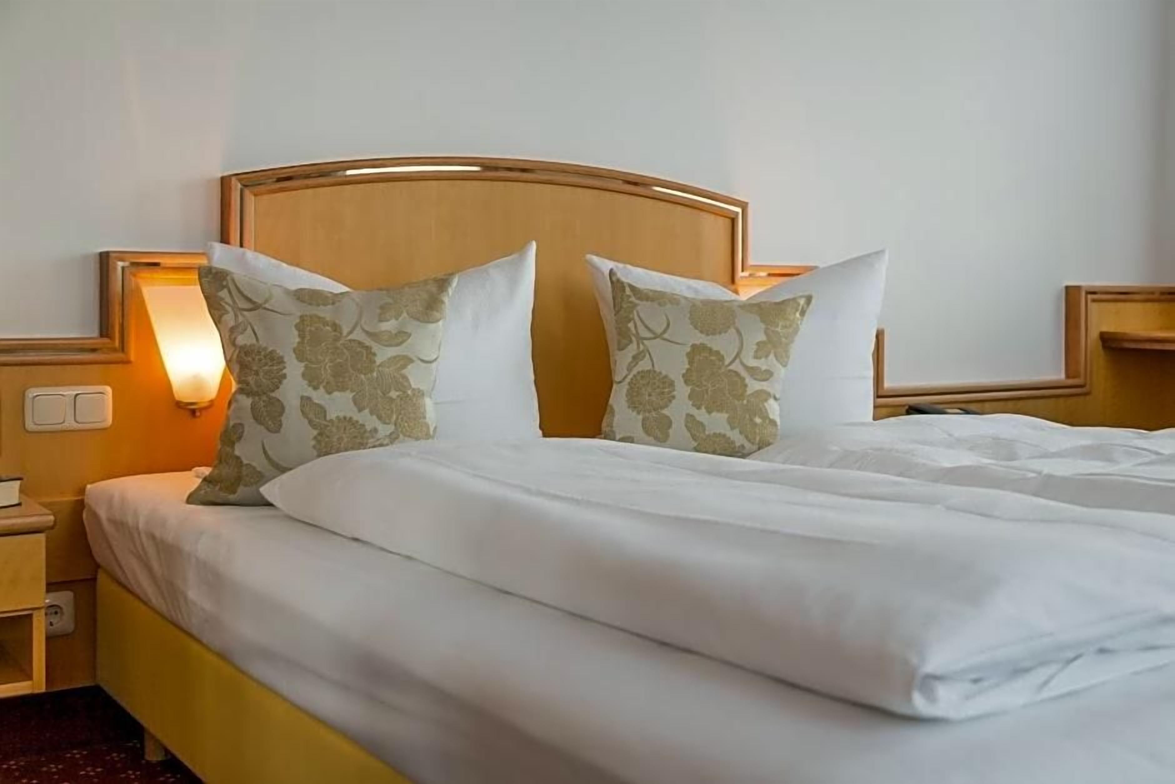 A neatly made bed with white bedding and two decorative pillows, a bedside table with a lamp, and a wall-mounted light fixture.