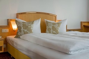 A neatly made bed with white bedding and two decorative pillows, a bedside table with a lamp, and a wall-mounted light fixture.