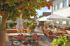 An outdoor seating area with wooden tables and chairs, surrounded by potted plants and a building with signage.