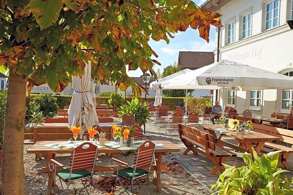 An outdoor seating area with wooden tables and chairs, surrounded by potted plants and a building with signage.