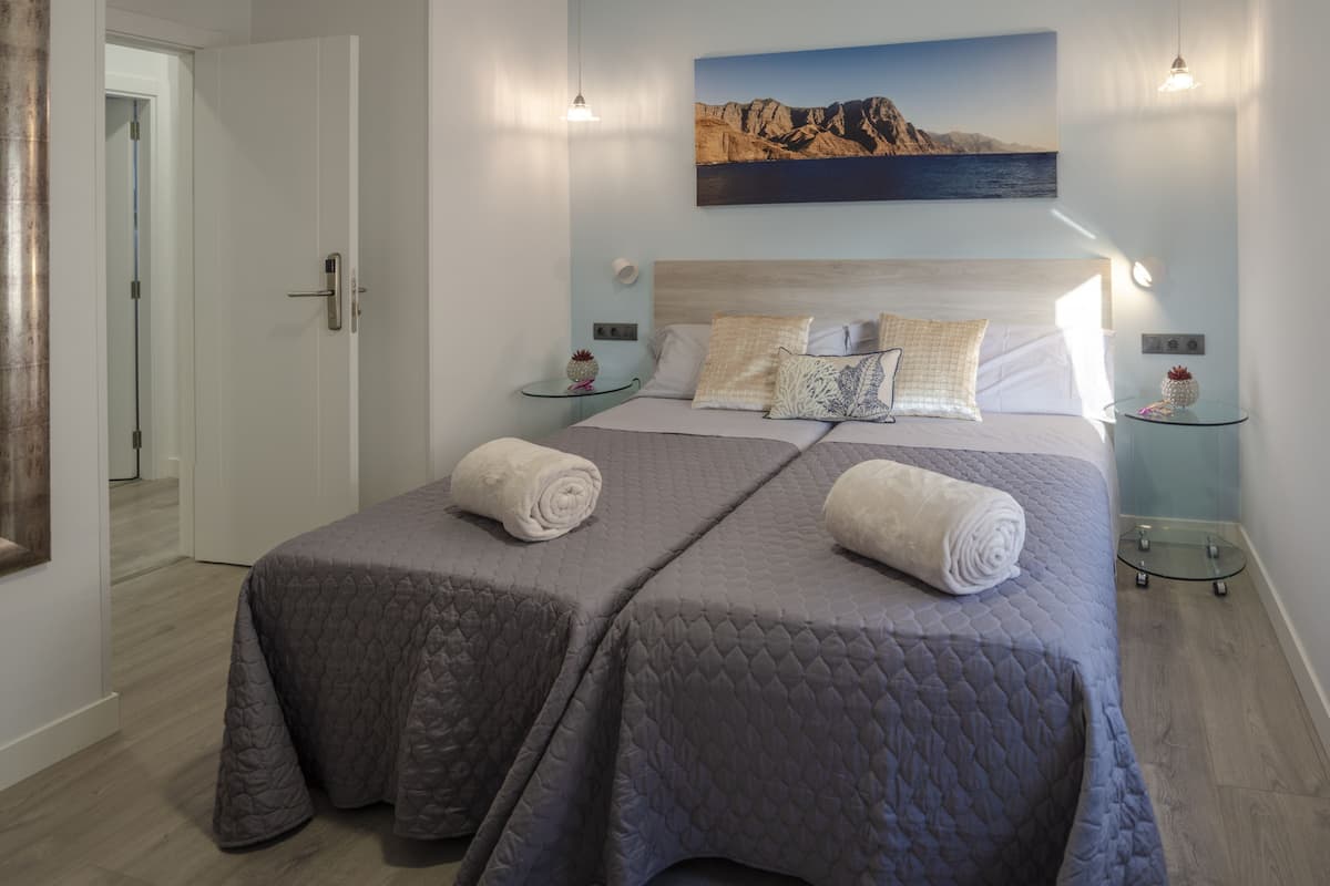 Un lit double avec des couvertures grises et des serviettes enroulées, une table de chevet avec une lampe, un tableau accroché au mur et une porte sur la gauche.