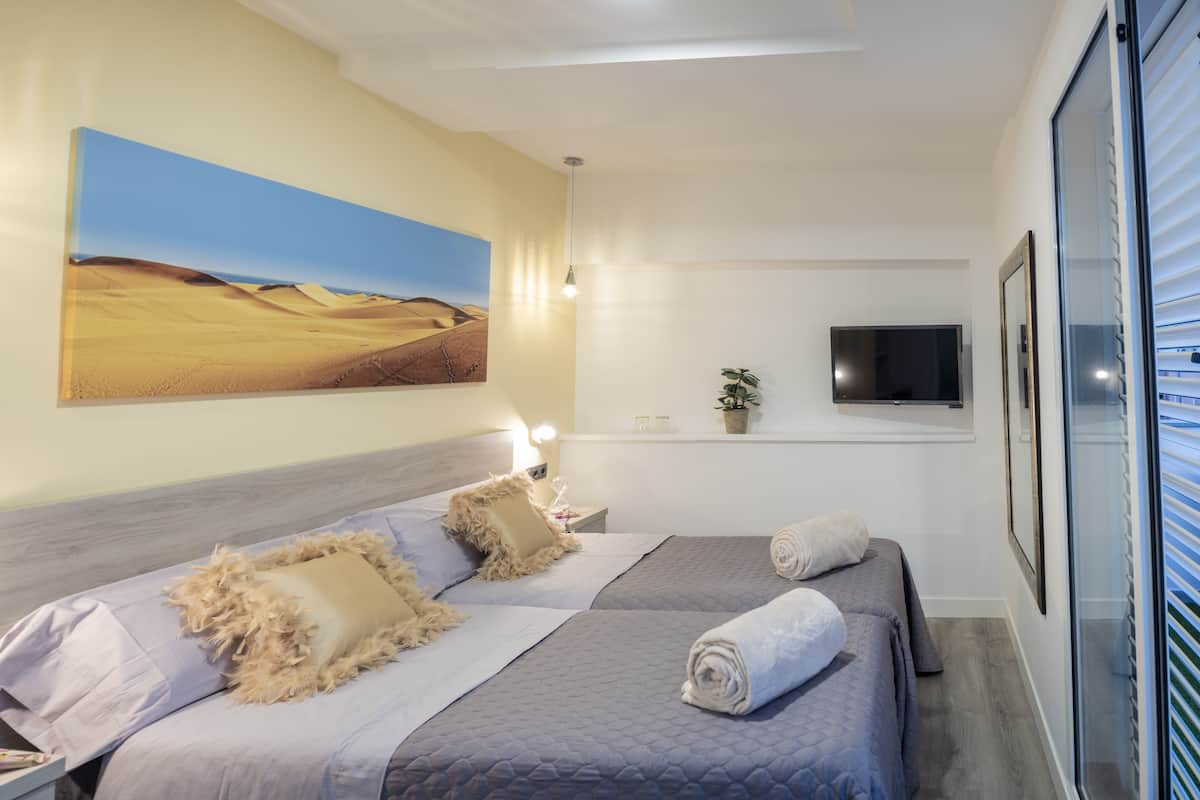 Une chambre d’hôtel avec deux lits, un grand tableau représentant un paysage désertique, un téléviseur à écran plat, une plante en pot et une fenêtre avec des stores.