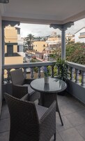 A balcony with wicker furniture and a glass table, overlooking a cityscape with buildings and a palm tree.