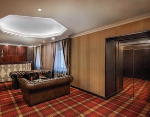 Habitación de hotel con sofá de cuero, alfombra escocesa, pared de paneles de madera y un diseño de techo moderno.