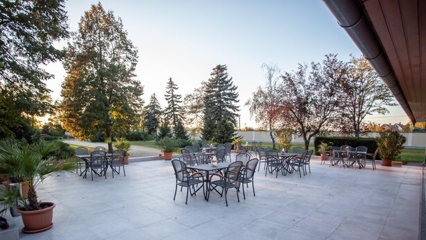 An outdoor patio with multiple tables and chairs, surrounded by trees and potted plants. - Arcanum Hotel (Bekescsaba)