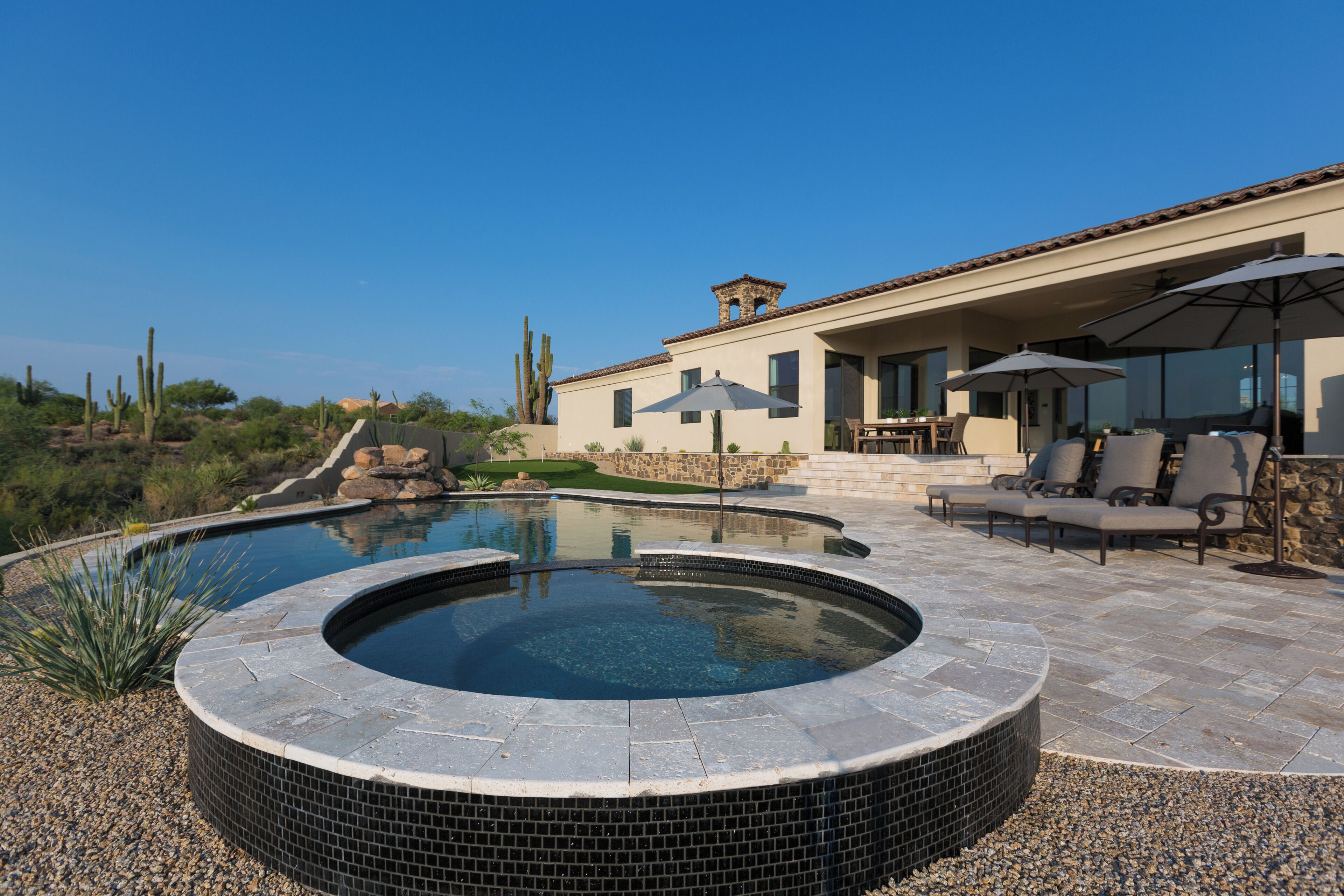 A hot tub with a tiled edge, a patio with chairs and umbrellas, and a house with a stone facade and large windows.