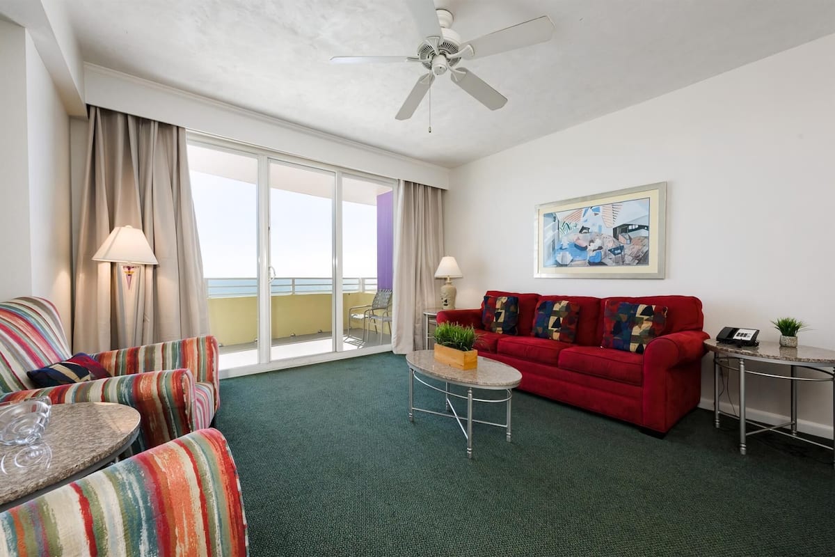 A hotel room with a red sofa, striped armchairs, a small round table with a plant, a ceiling fan, and a large window with curtains.