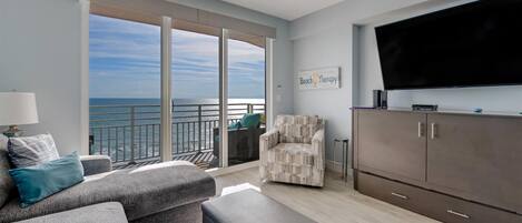 A modern living room with a grey sofa, a patterned armchair, a flat-screen TV, and a large window overlooking the ocean.