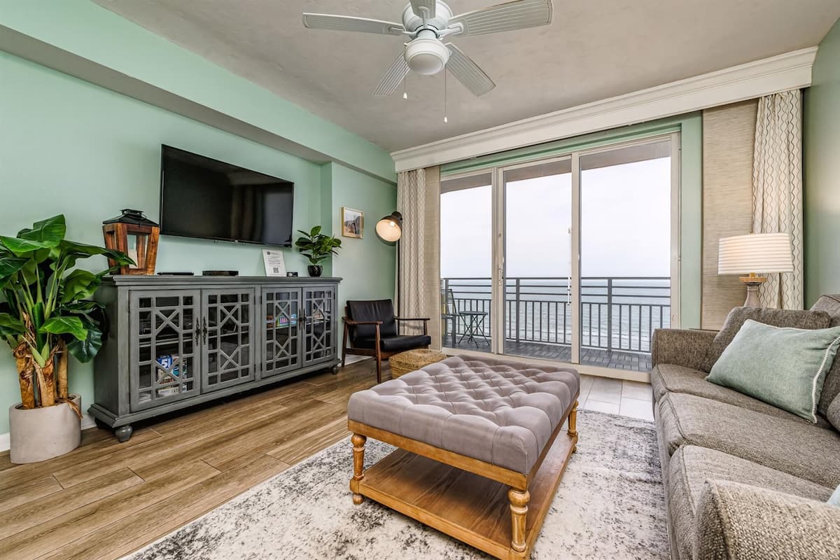 A living room with a large flat-screen TV, a grey cabinet, a wooden bench, a brown sofa, and a balcony with a view.
