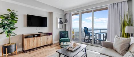 Een moderne woonkamer met een flatscreen televisie, een houten kast, een groene fauteuil, een beige bank en een balkon met uitzicht op zee.