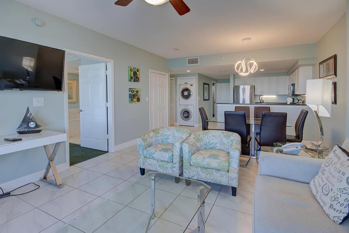 A modern living room with a sofa, two armchairs, a glass coffee table, and a TV. There is a kitchen area with a dining table and chairs, and a laundry room with a washing machine and dryer.