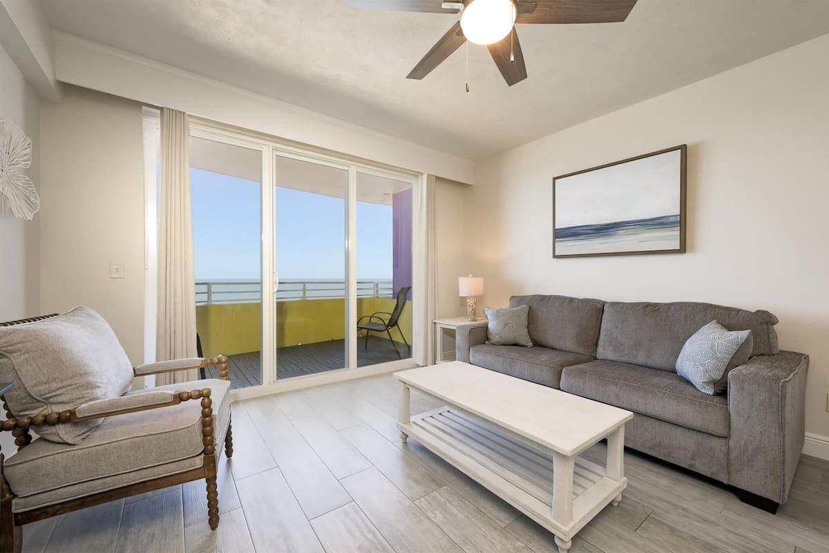A living room with a grey sofa, a wooden chair, a white coffee table, and a ceiling fan. There is a large window with a view of the ocean.