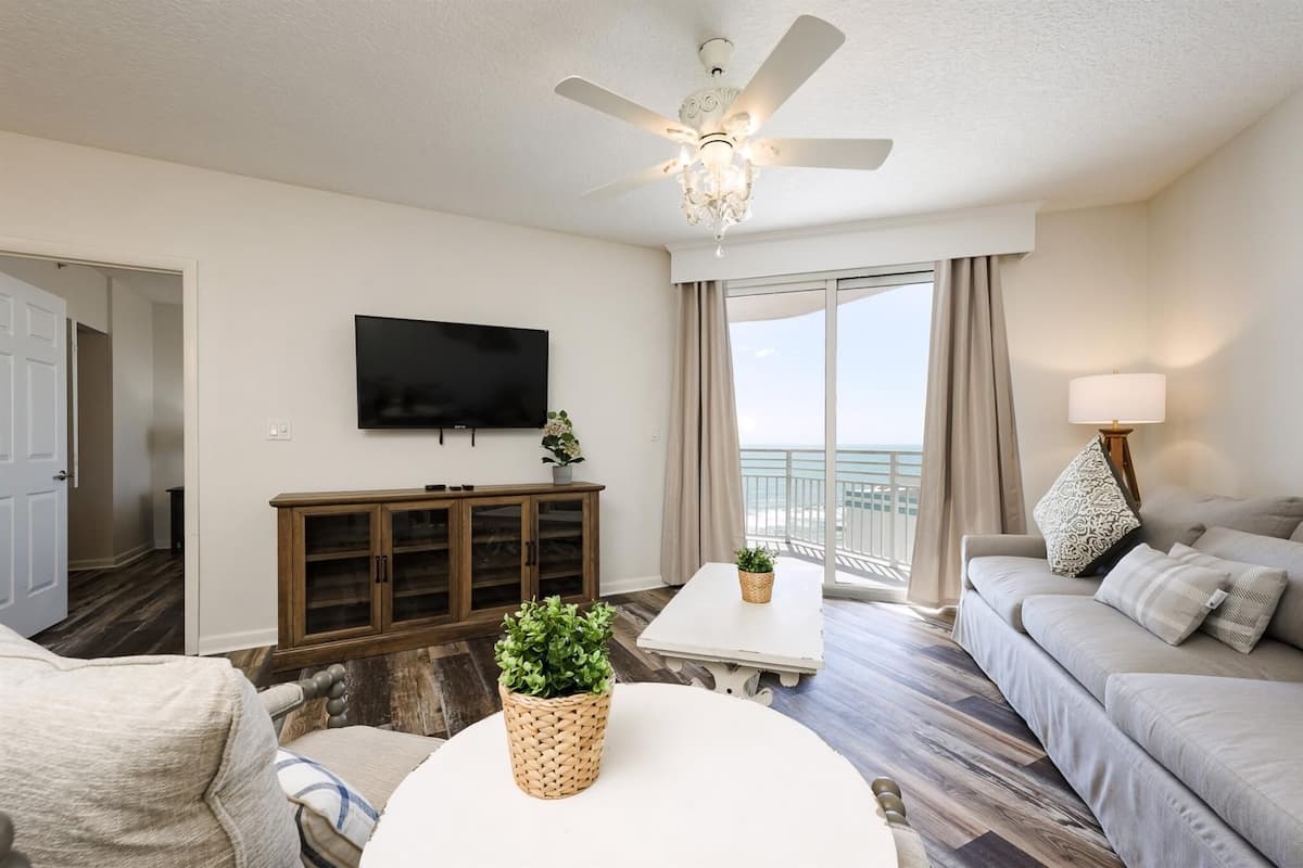 A modern living room with a ceiling fan, a flat-screen TV, a sofa, a coffee table, and a view of the ocean through large windows.