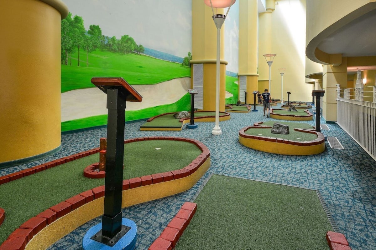A miniature golf course with multiple holes, each with a flag, surrounded by a brick border and green turf.