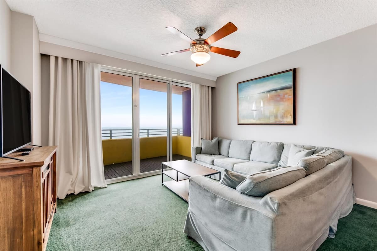 A modern living room with a grey sofa, a wooden coffee table, a ceiling fan, and a large window with curtains.