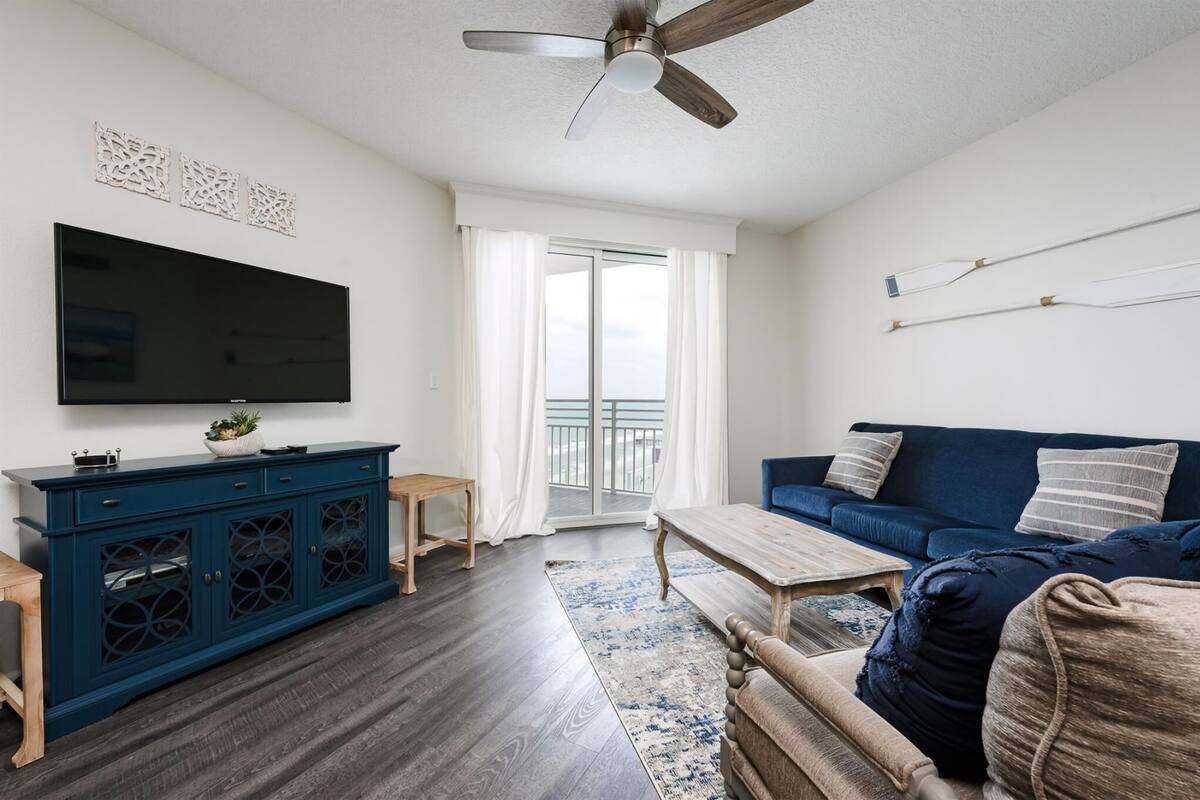 A modern living room with a dark blue sofa, a flat-screen TV, a wooden coffee table, and a ceiling fan.