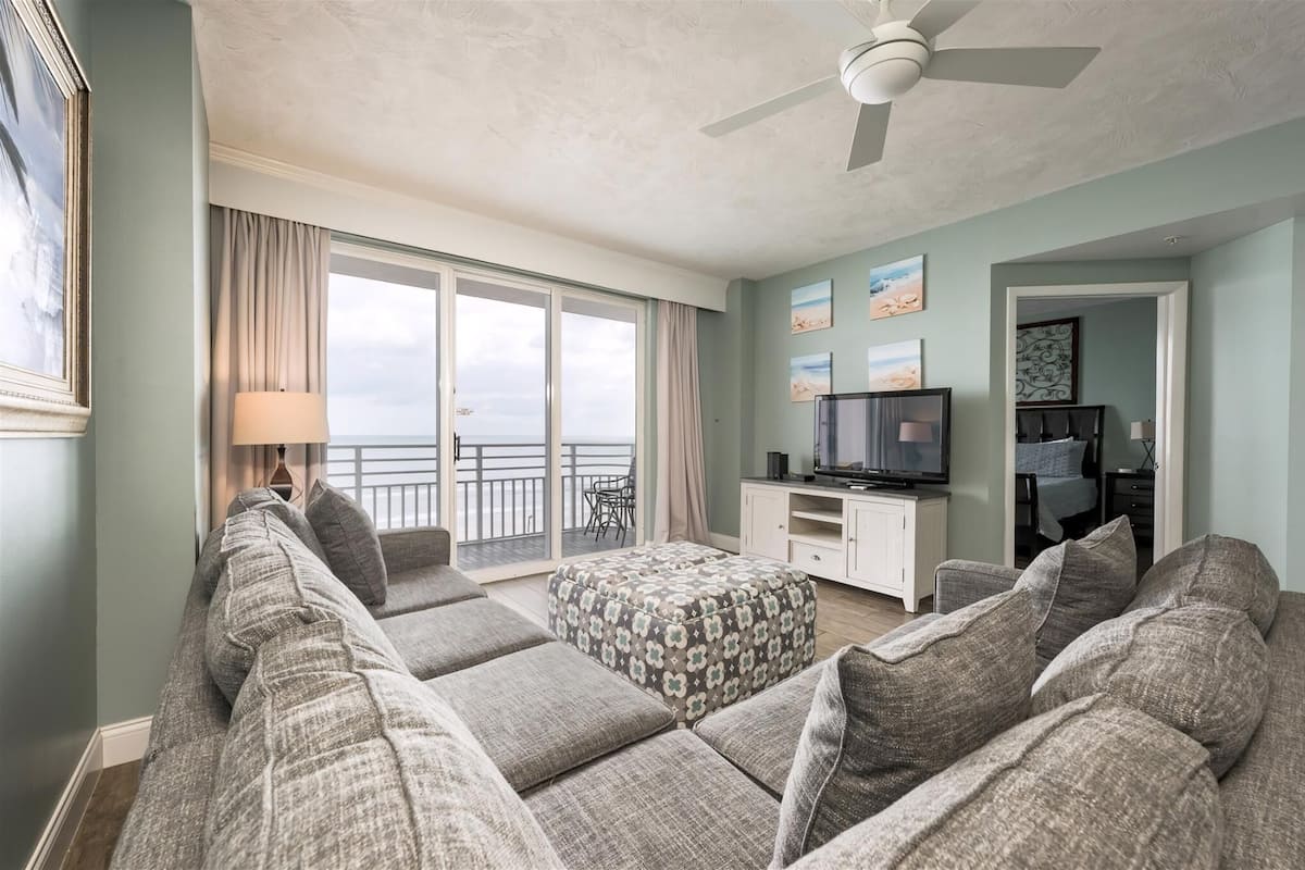 A spacious living room with a large sofa set, a television, and a balcony with a view.