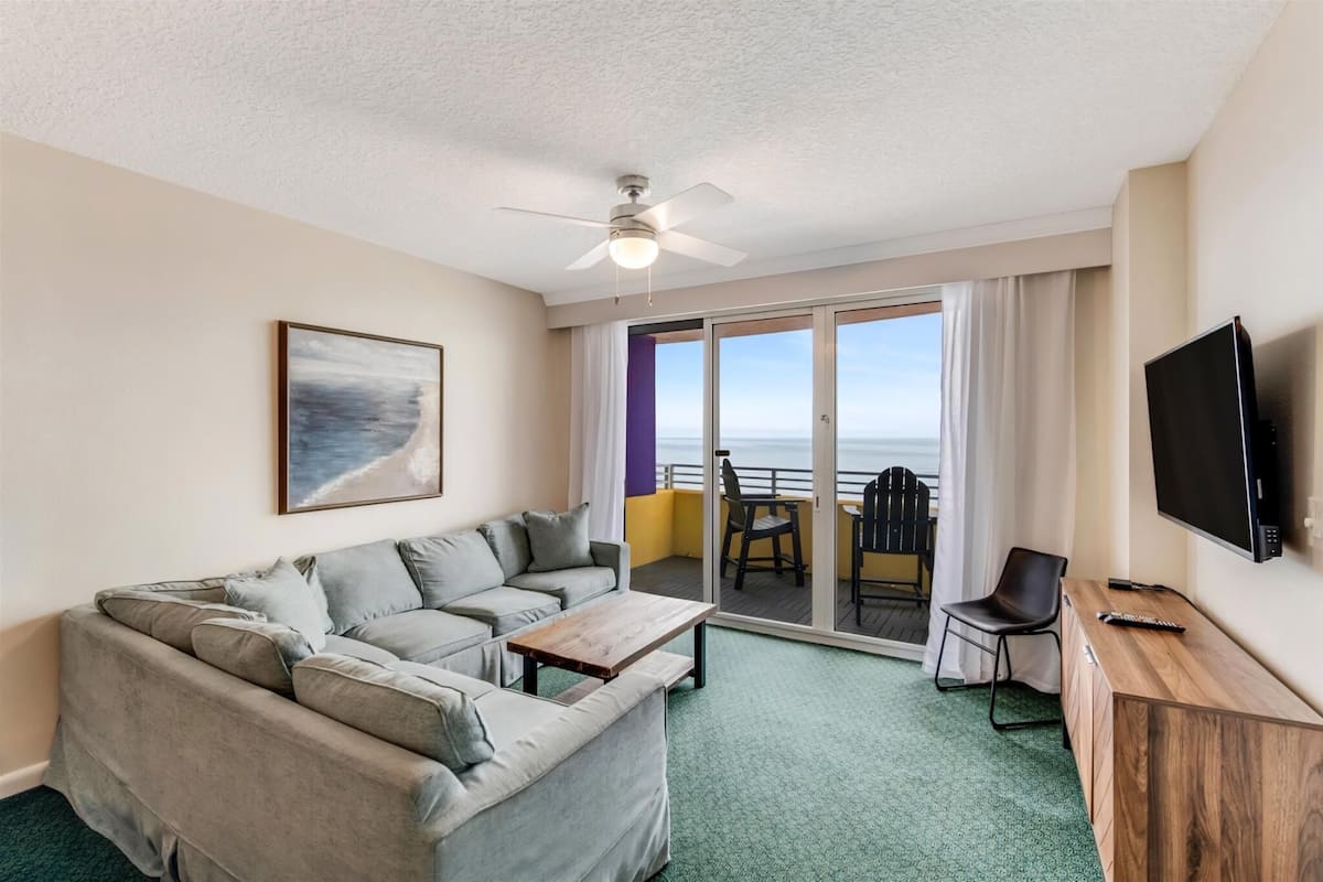A hotel room with a sofa, a wooden coffee table, a TV, and a balcony with a view of the ocean.