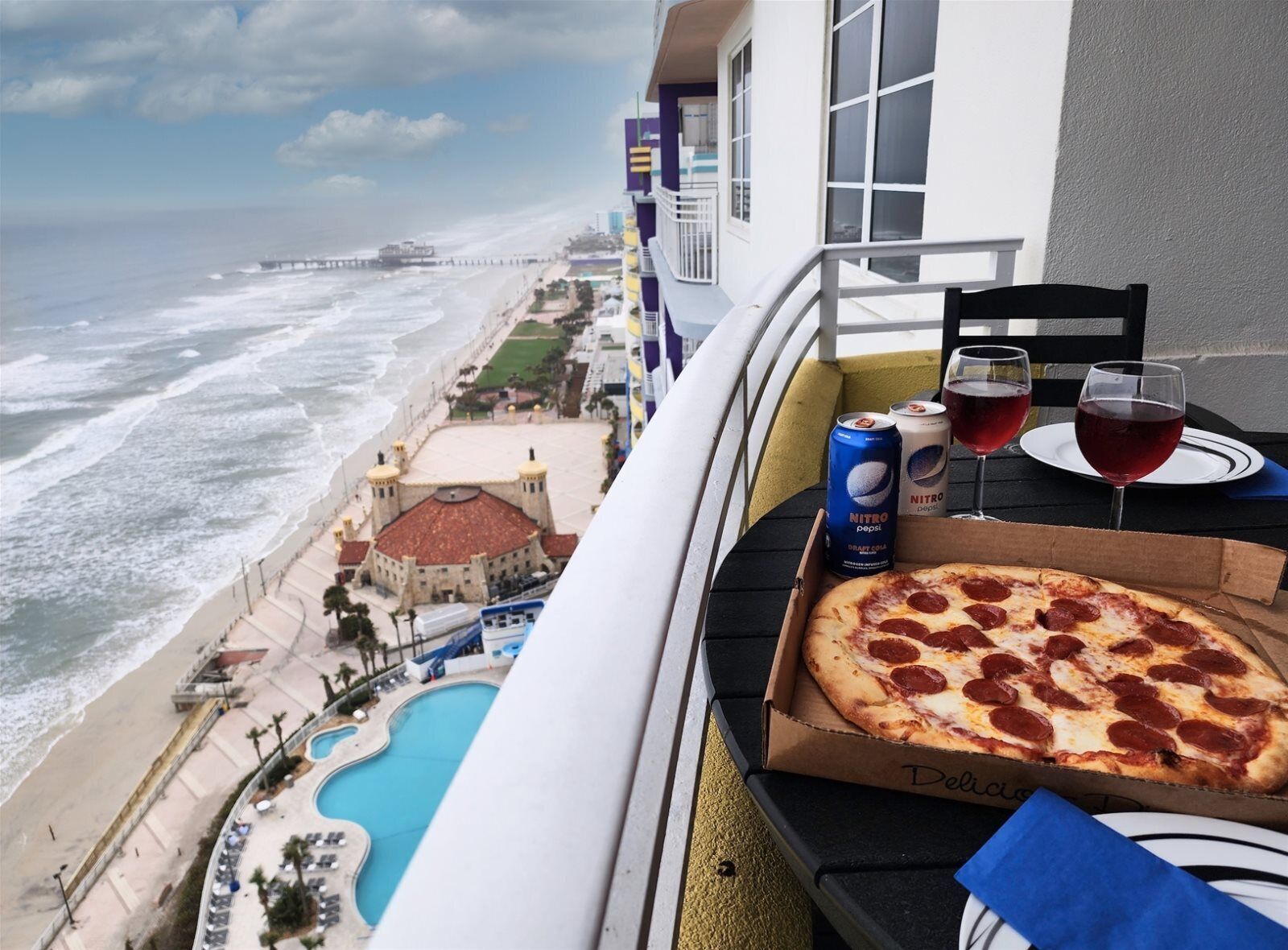 Un balcon avec un carton de pizza, deux canettes de Pepsi, deux verres de vin rouge et une table.