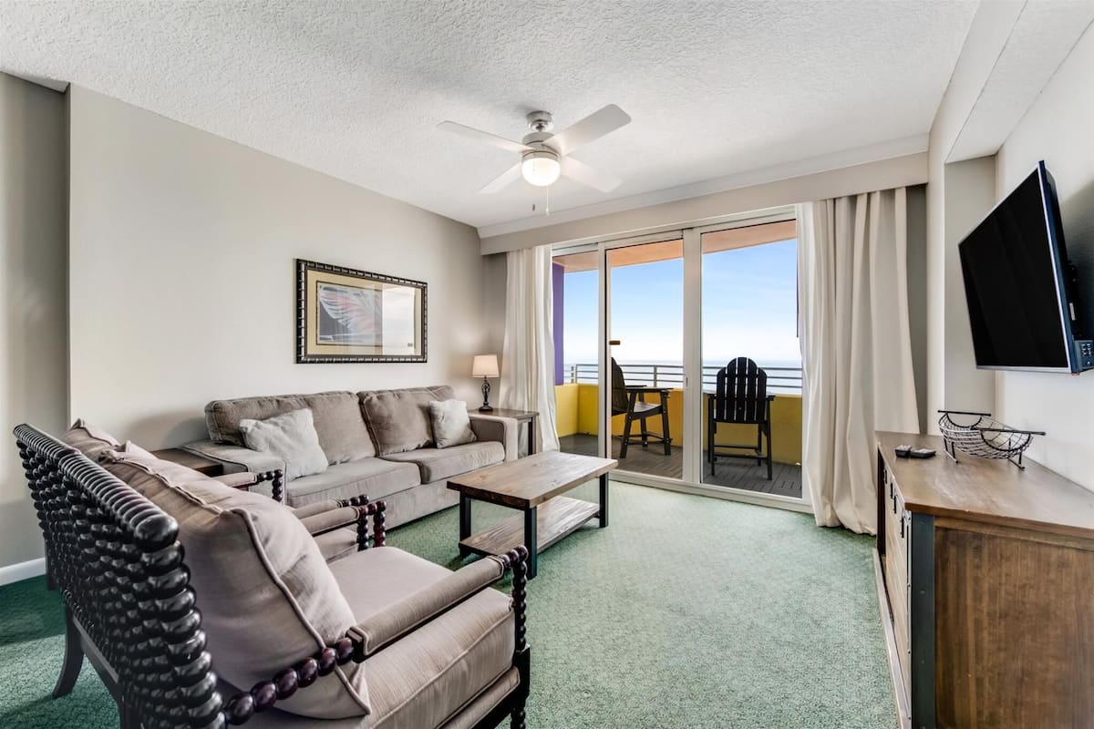 A hotel room with a sofa, armchair, and a small table. There is a TV mounted on the wall and a balcony with a view.