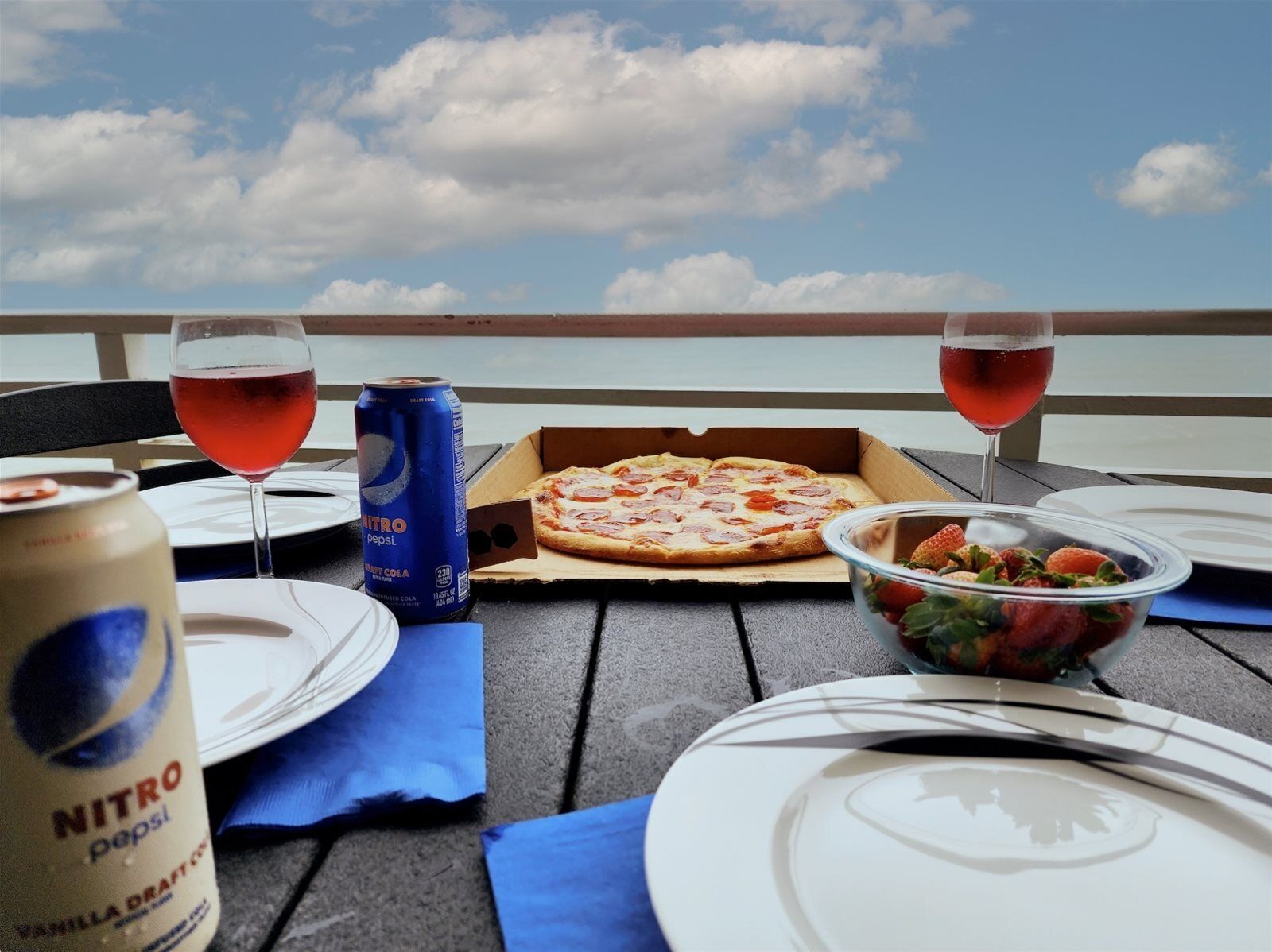 A table set with a pizza box, two glasses of red drink, a can of Pepsi, a bowl of strawberries, and a bottle of Nitro Pepsi.