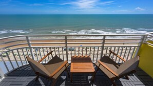 Un balcon avec des fauteuils en osier et une table en bois, donnant sur une plage où l’on peut admirer les vagues et un ciel dégagé.