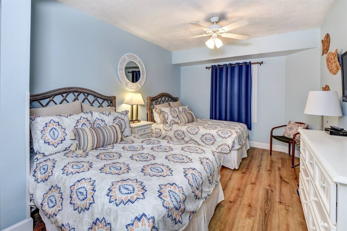 A bedroom with two beds, a ceiling fan, a mirror, a dresser, and a window with blue curtains.