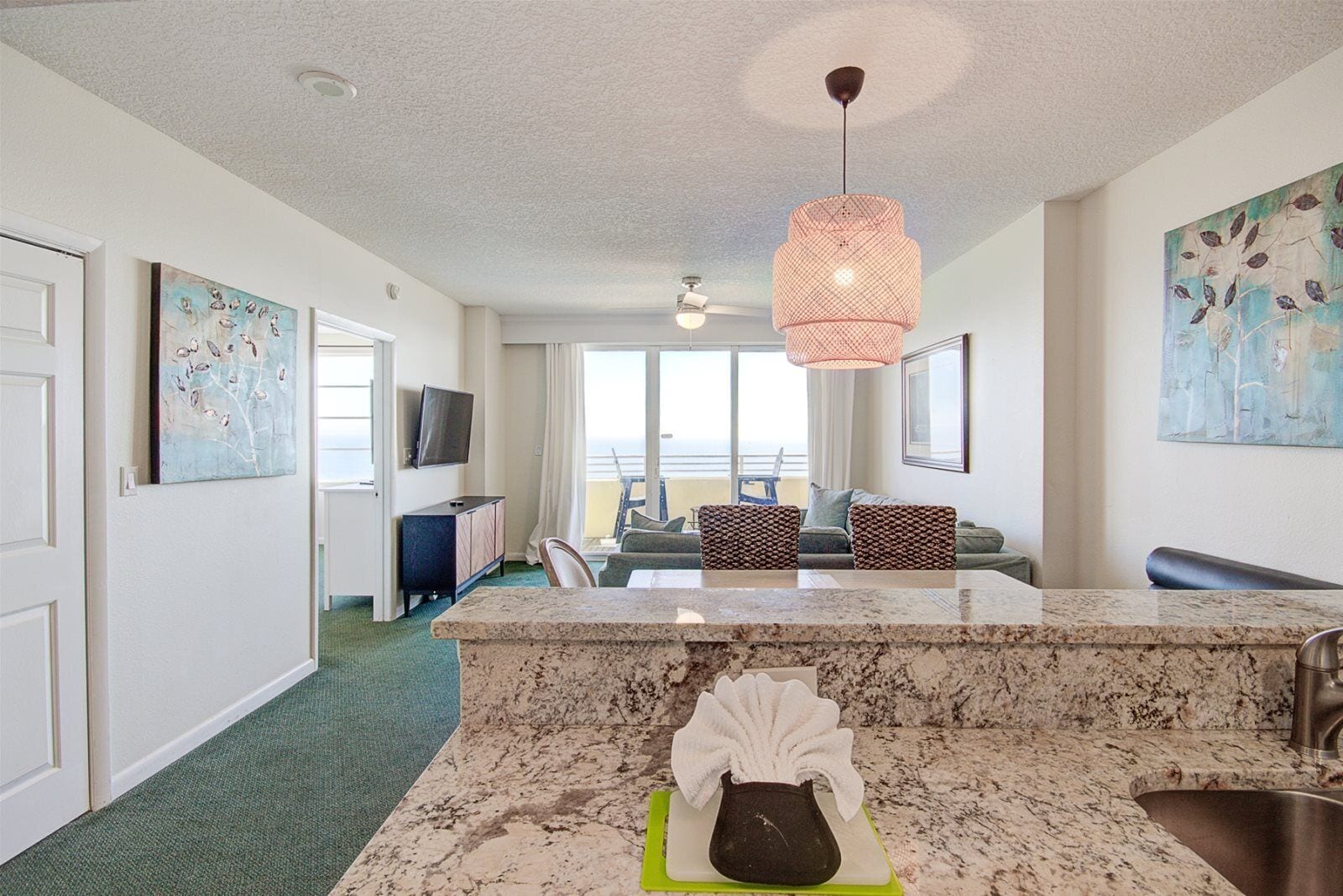 A modern hotel room with a marble kitchen island, a dining area with a table and chairs, a large window with a view, and artwork on the walls.