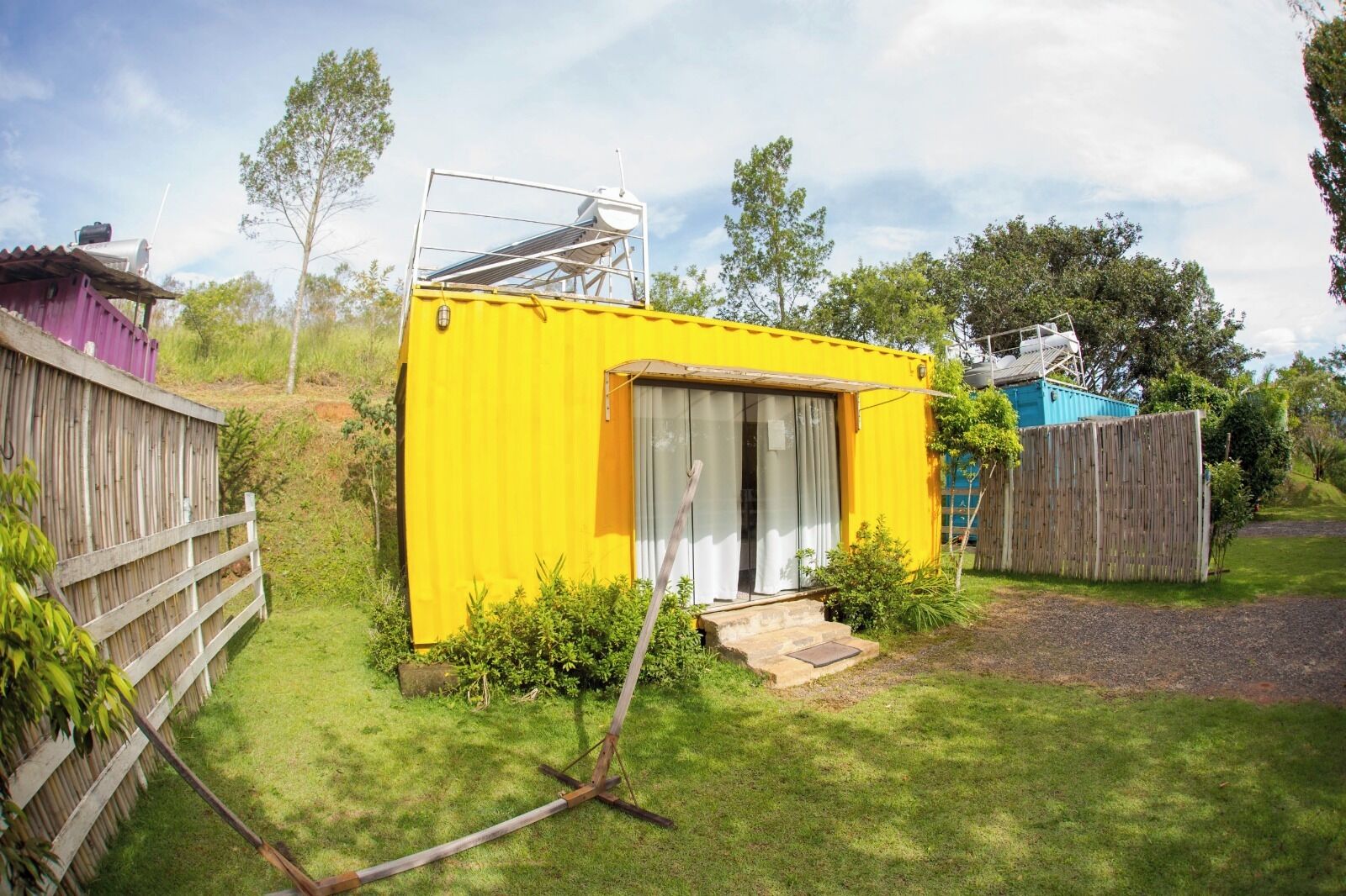 A small, yellow, single-story building with a sliding glass door, surrounded by a wooden fence and greenery.
