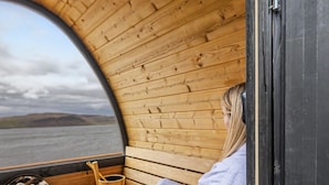 A wooden cabin with a bench and a bucket of stones, a window with a view of a body of water and hills, and a person sitting on the bench.