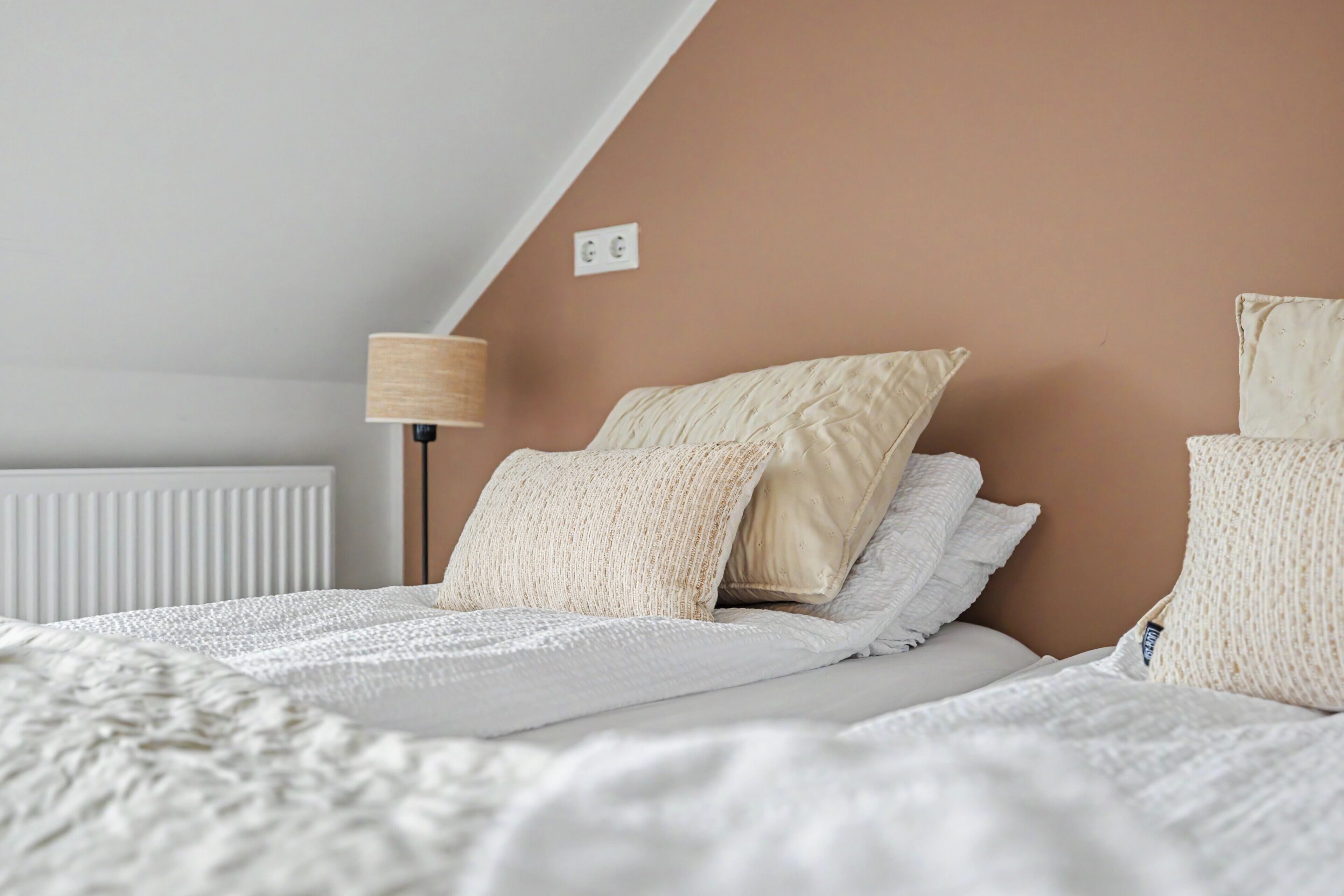 Un dormitorio con techo inclinado, un radiador, una lámpara y una cama con muchas almohadas y una colcha blanca.