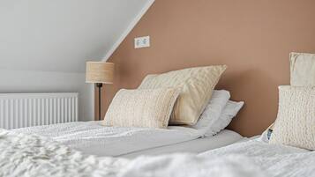Un dormitorio con techo inclinado, un radiador, una lámpara y una cama con muchas almohadas y una colcha blanca.