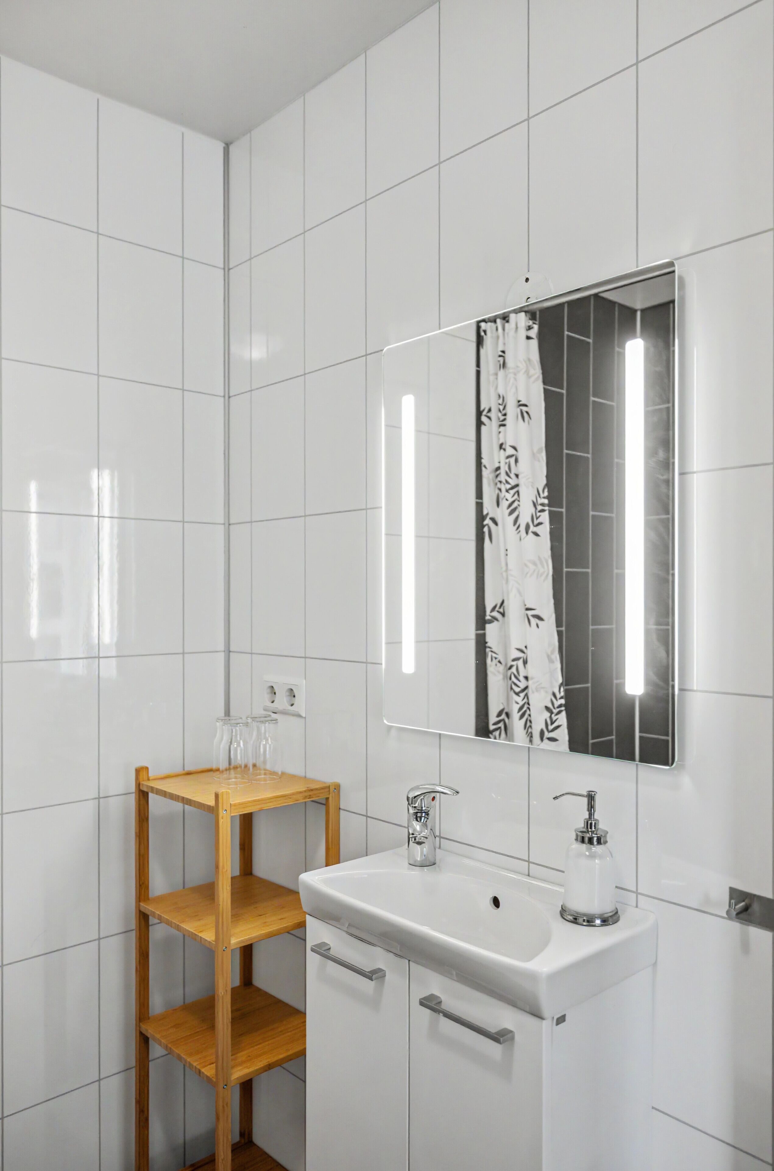 Bany amb parets revestides de rajoles blanques, una prestatgeria de fusta, un lavabo amb mirall i una cortina de banyera.