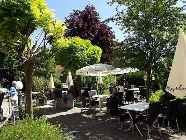An outdoor seating area with tables and umbrellas, surrounded by trees and greenery.