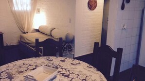 A small, cozy room with a bed, a table with a floral tablecloth, and a wall with a decorative plate and two hanging spoons.
