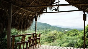 A wooden deck with chairs, a thatched roof, and a view of a green landscape with hills. - Hotel Beyonda (Santa Marta)
