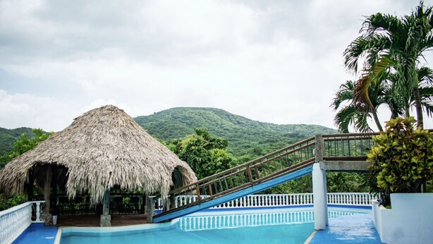 A pool area with a thatched-roof structure, a wooden slide, and a view of a lush green mountain. - Hotel Beyonda (Santa Marta)