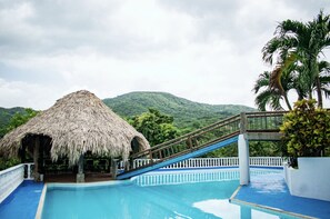 A pool area with a thatched-roof structure, a wooden slide, and a view of a lush green mountain. - Hotel Beyonda (Santa Marta)