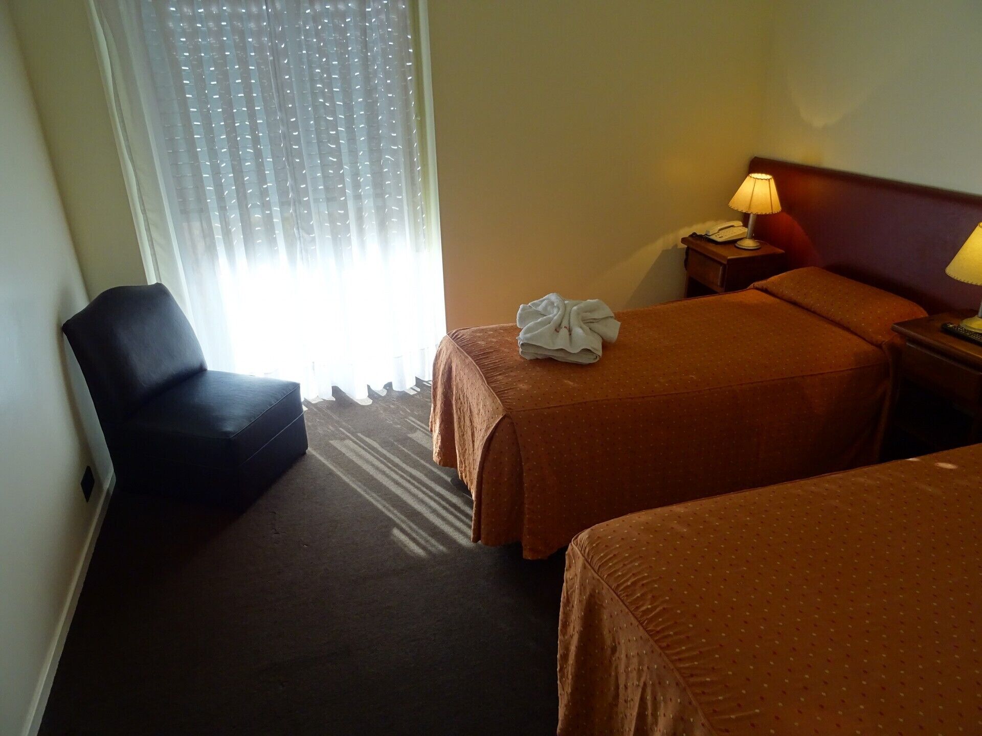 A hotel room with two beds, a chair, a nightstand with a lamp, and a window with sheer curtains.
