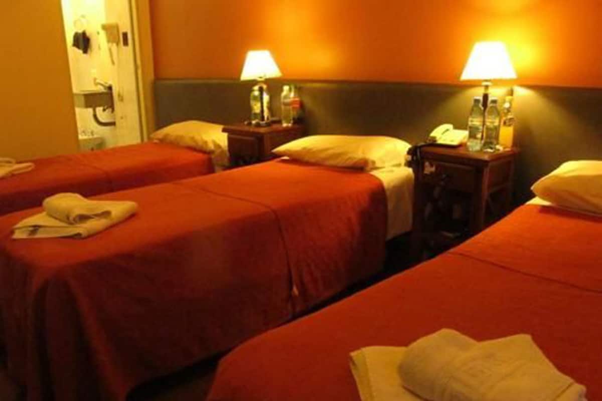 A hotel room with three beds, each with a red bedspread and white pillows. There are two bedside tables with lamps and bottles.