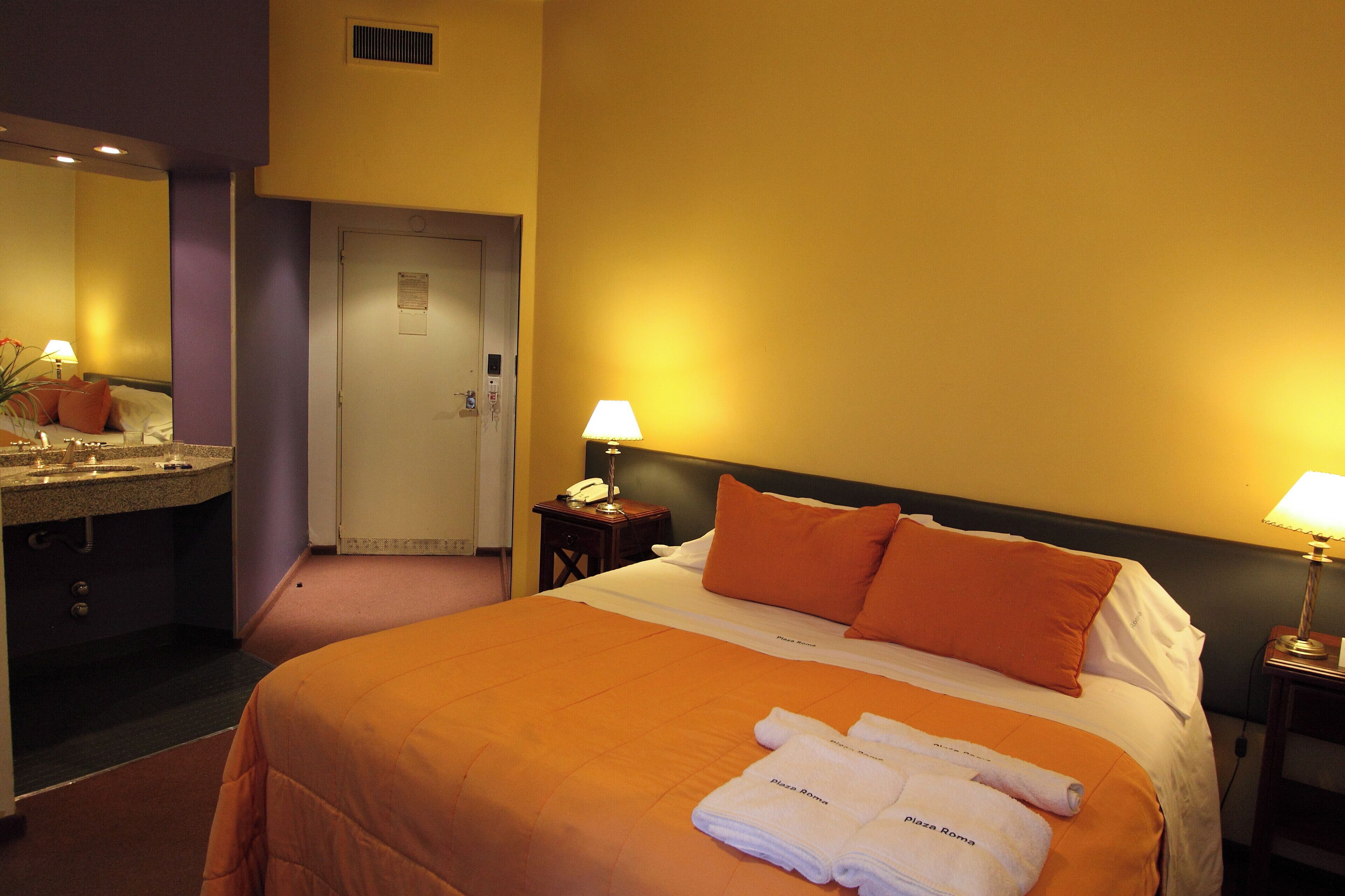 A hotel room with a bed, two bedside lamps, a nightstand, a bathroom with a sink and mirror, and a door.