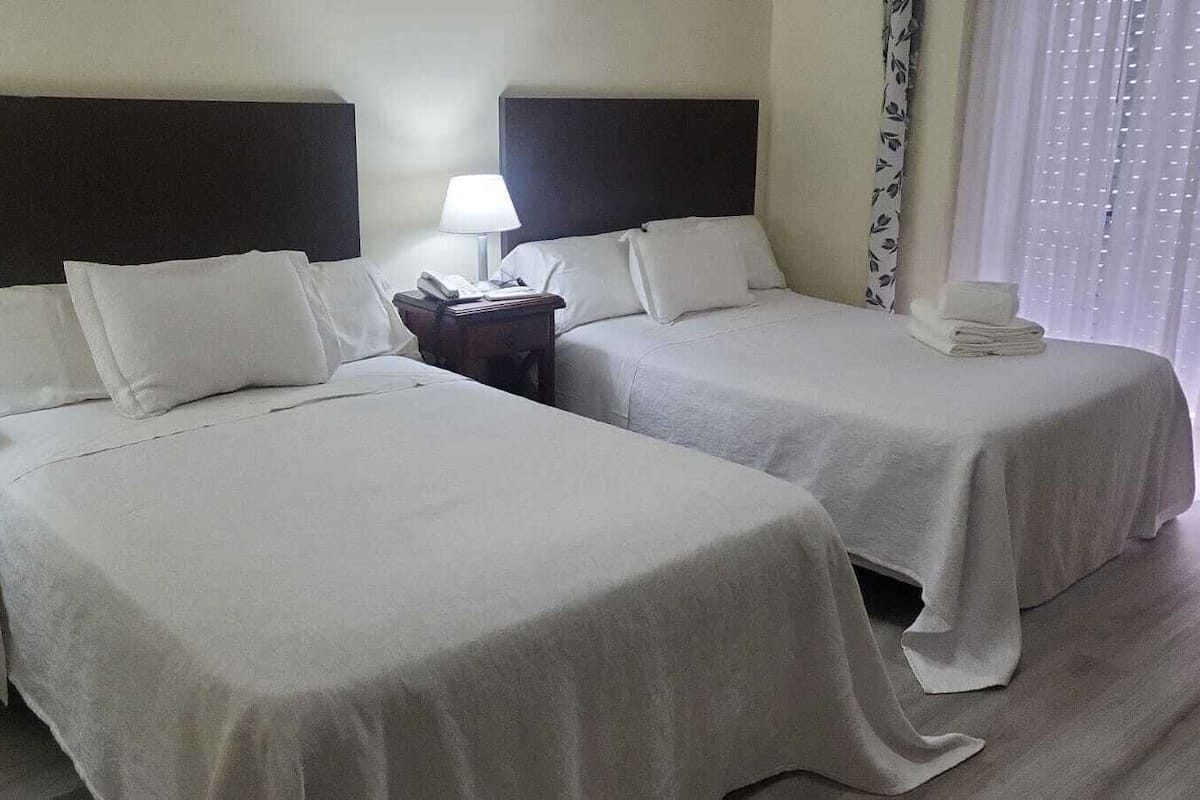 A hotel room with two beds, a nightstand with a lamp, and a window with curtains.