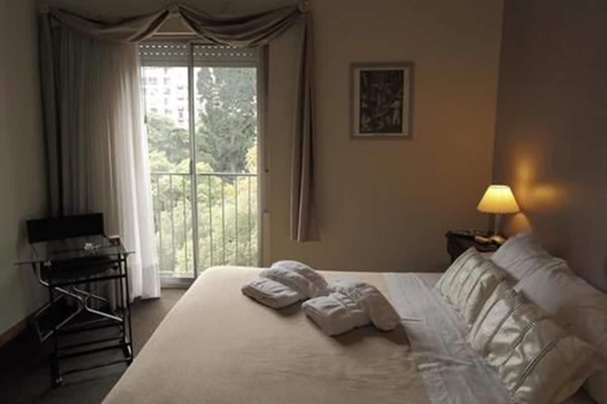 A hotel room with a bed, a desk, a chair, a lamp, and a window with curtains.