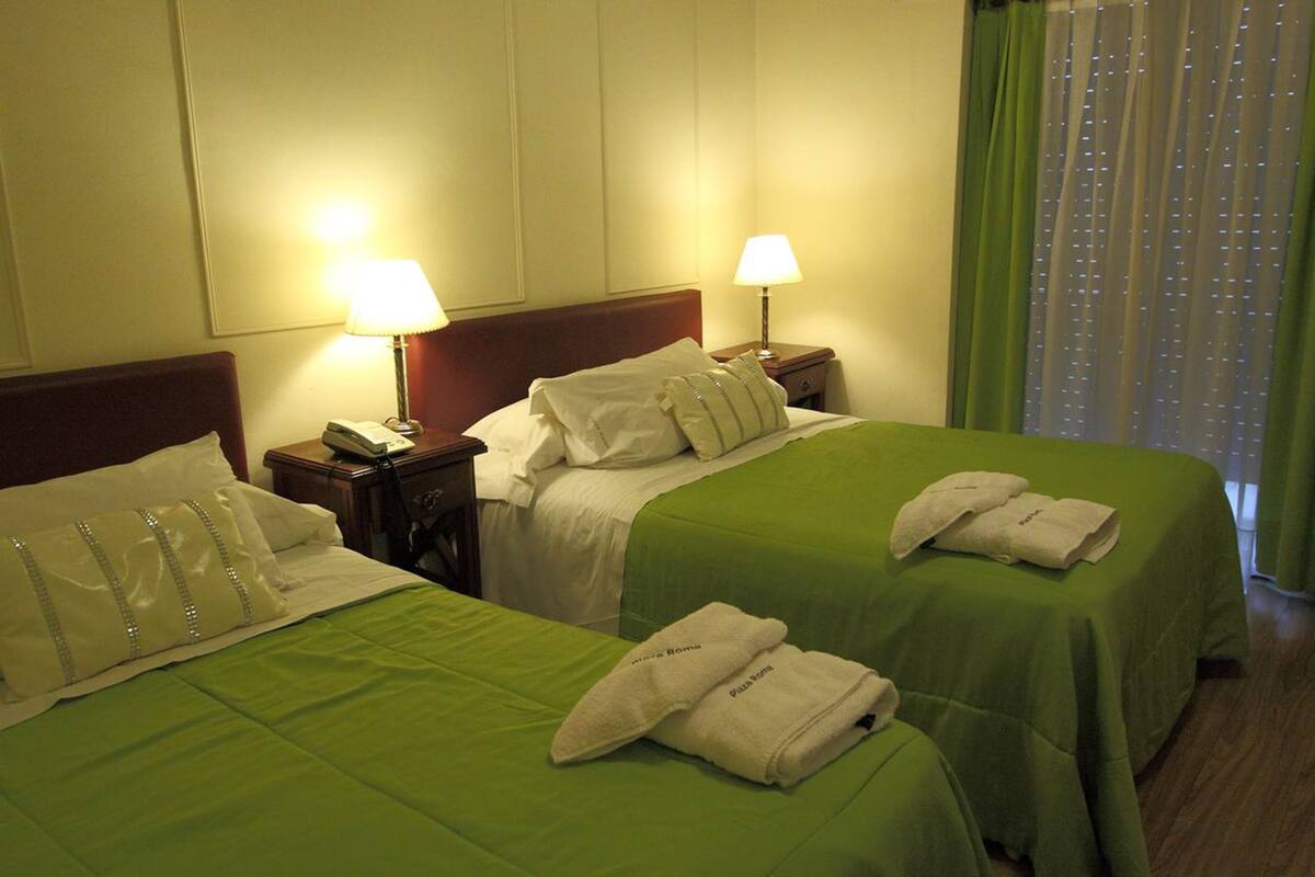 A hotel room with two beds, each with a green bedspread and white linens. There are two bedside lamps, a wooden nightstand, and a window with green curtains.