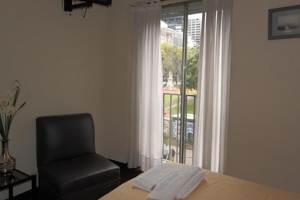Une chambre d’hôtel avec un lit, une chaise noire, une petite table avec un vase et une fenêtre donnant sur un parc et des immeubles.